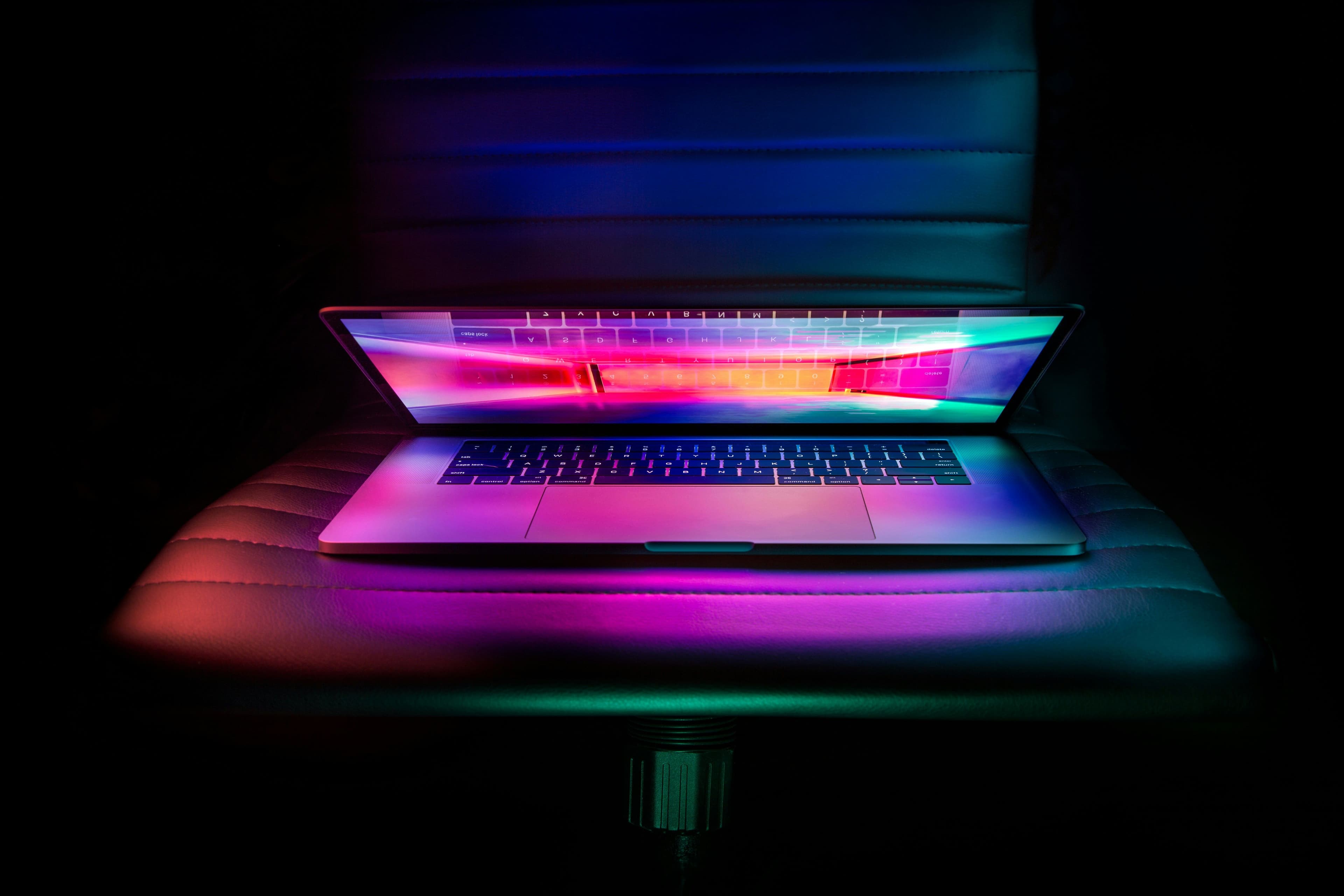 Ein geöffneter Laptop liegt auf einem leeren Stuhl und wird von farbigem Licht angestrahlt.