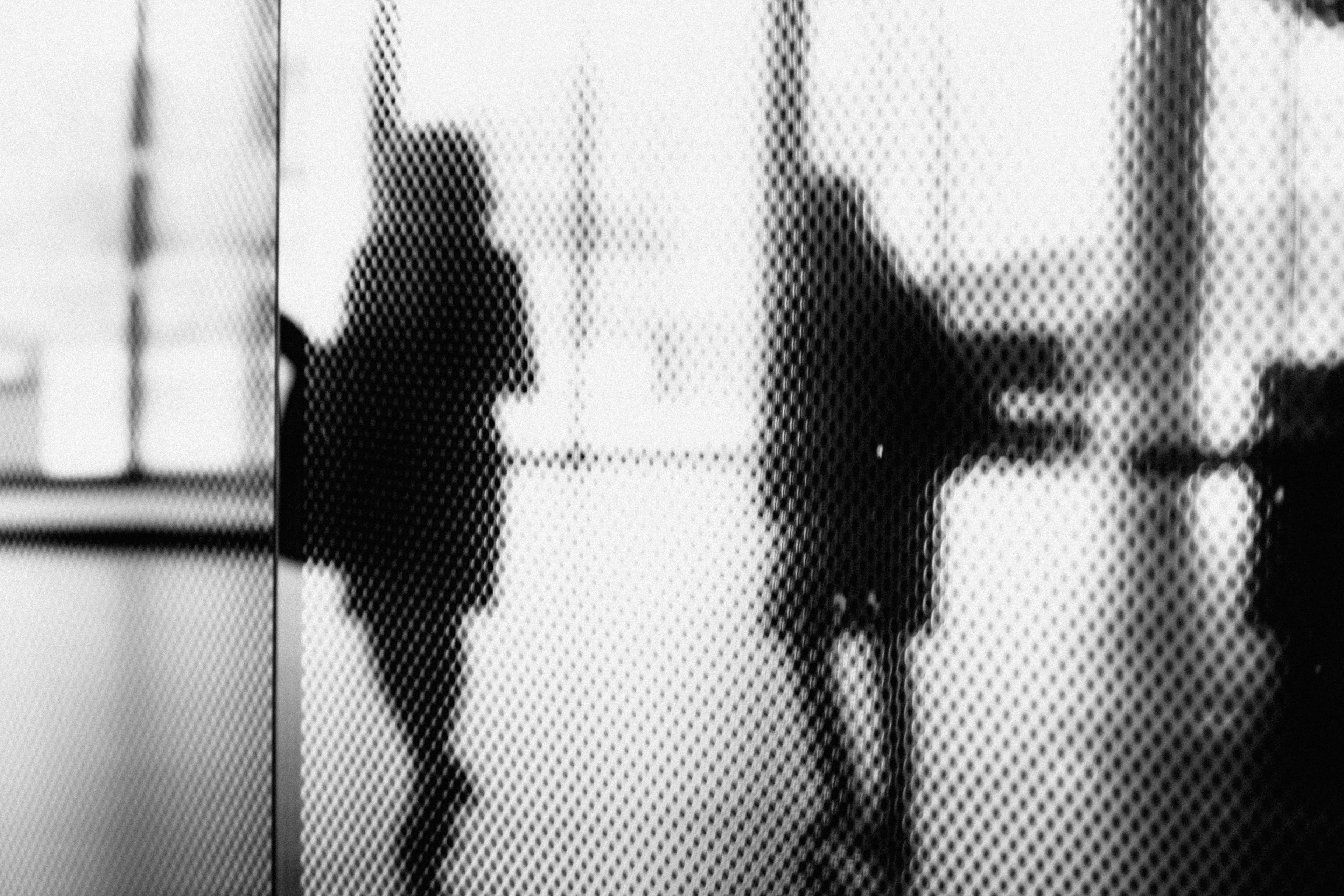 Schattenhafte Person hinter perforierter Glas- oder Metallfläche als Bild für geschützte Datenräume, Kontrolle und eingeschränkte Sichtbarkeit.