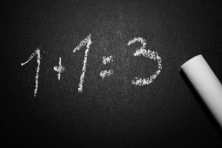 Mit Kreide geschriebene Rechnung „1 + 1 = 3“ auf dunklem Untergrund neben einem Kreidestück – Symbol für fehlerhafte Selbsteinschätzung.