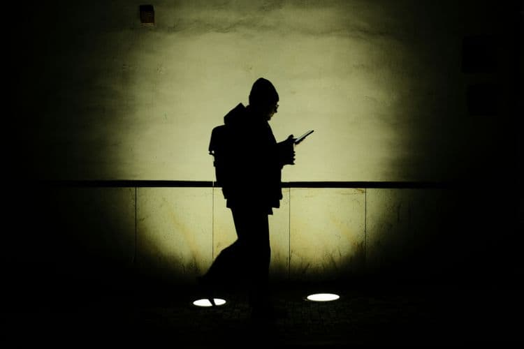 Silhouette einer Person mit Rucksack, die in dunkler Umgebung auf ein Smartphone blickt. Die Person ist von einem hellen Lichtkegel an der Wand hinter ihr umrahmt.