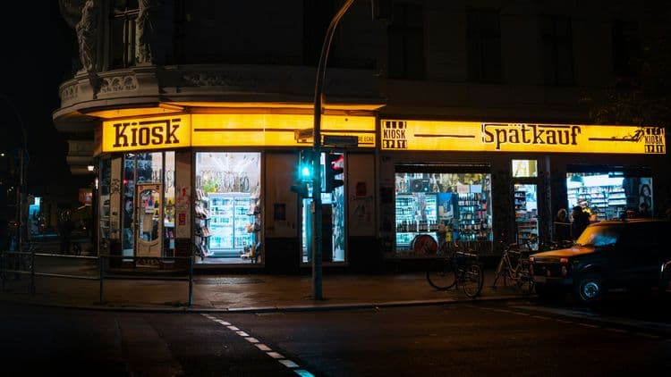 Nächtliche Außenansicht eines beleuchteten Berliner Spätkaufs mit Kiosk-Schriftzug, Schaufenstern, Getränkeregalen und Fahrrädern vor dem Laden.