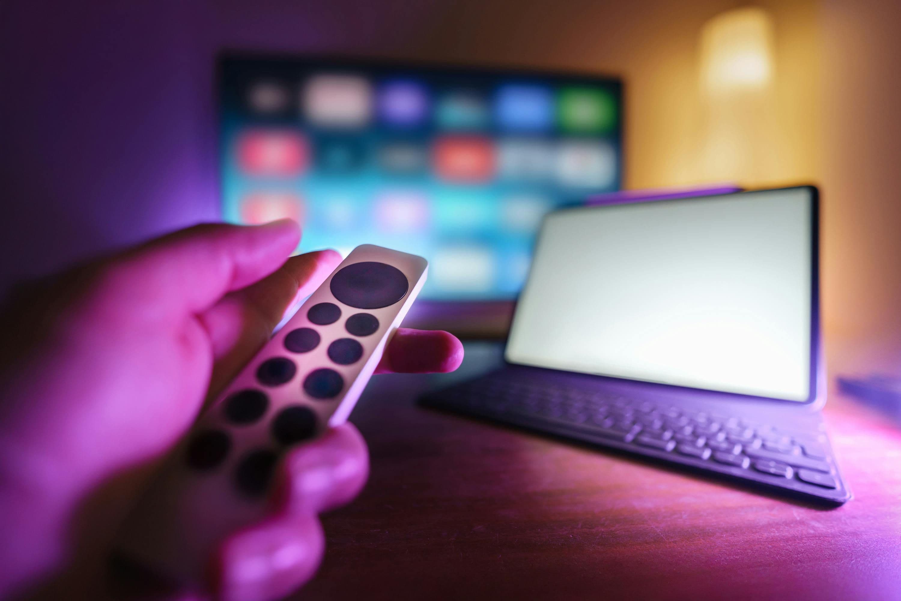 Hand hält eine Fernbedienung vor einem unscharfen Streaming-Bildschirm, daneben steht ein Tablet.
